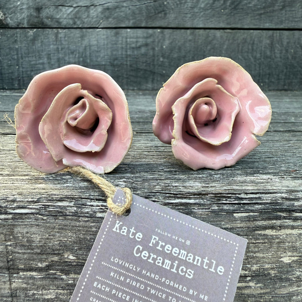 Two pink ceramic roses on a wooden surface with a tag from Kate Freemantle Ceramics.